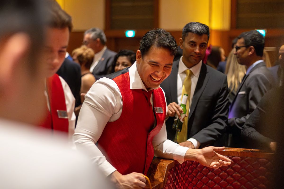 A dealer in a red vest laughing with guests around a casino table, photographed by Dario Hudon